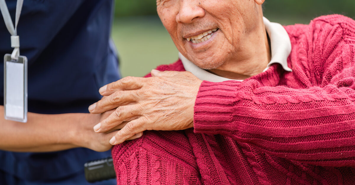 Elderly man smiling joyfully as a caregiver gently places a hand on his shoulder outdoors