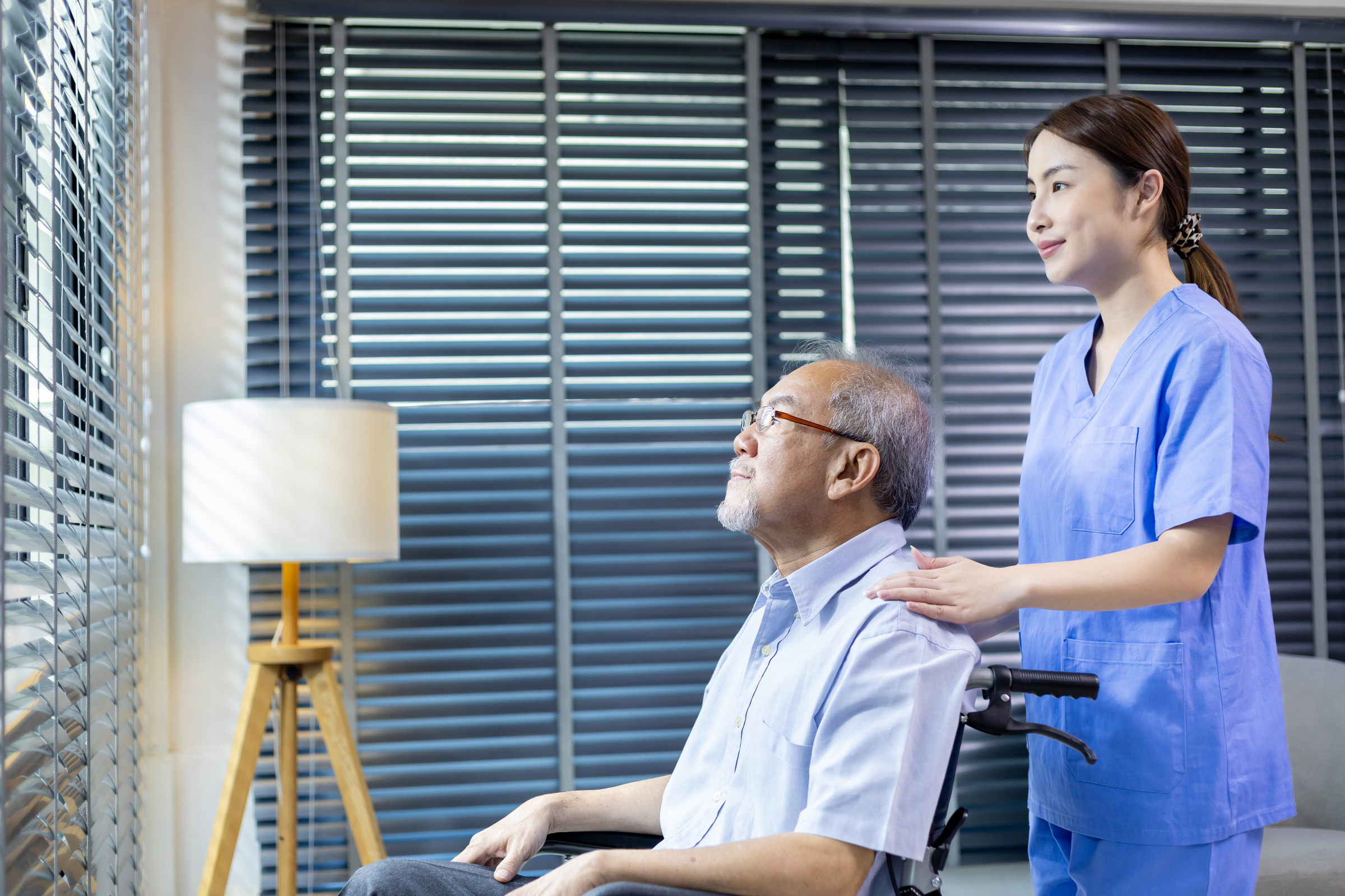 Asian senior man sitting in wheelchair at hospital looking out t Comfort Home Care Services caregiver assisting an elderly man in a wheelchair while looking out the window at home