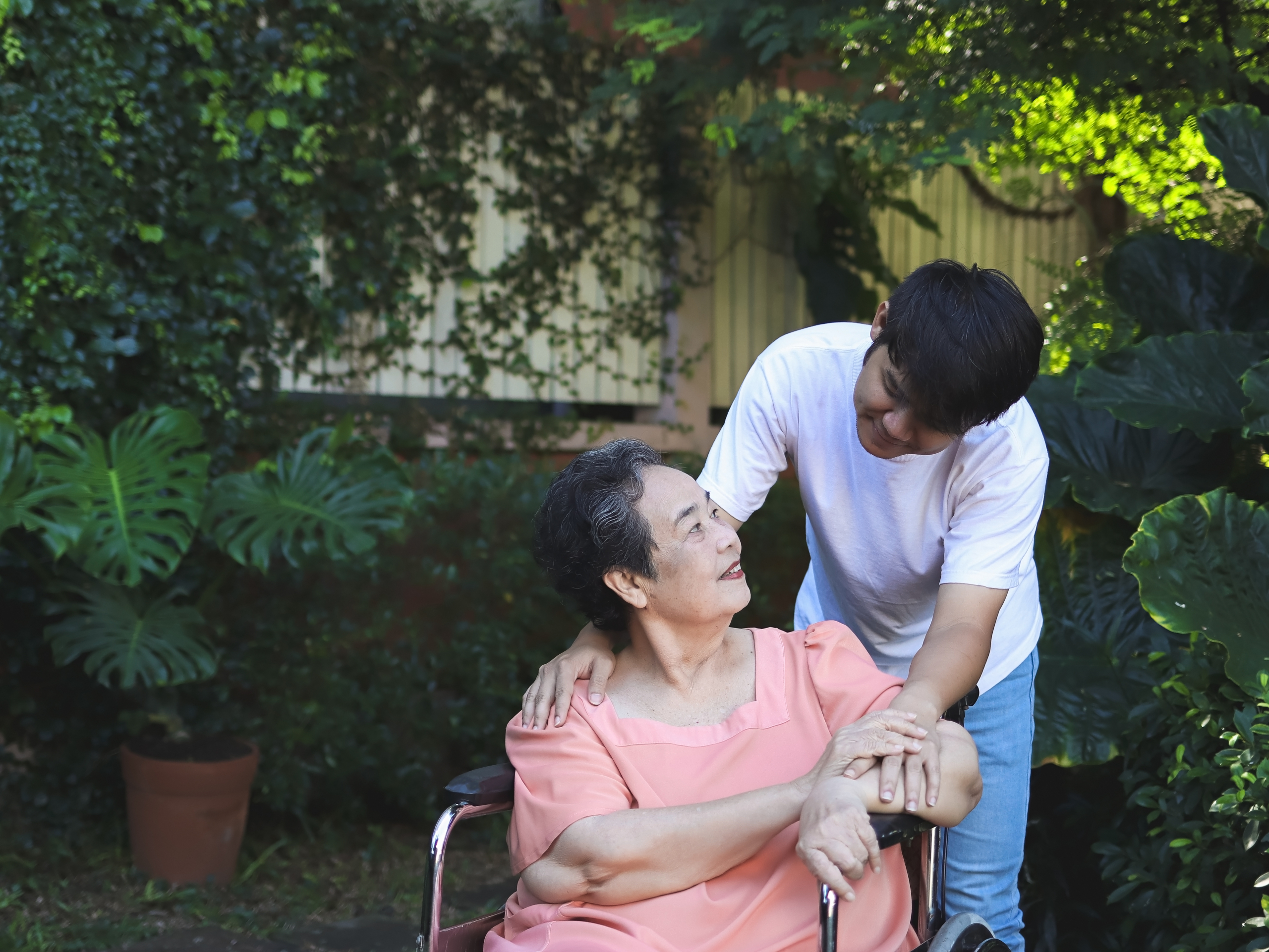 Happy Asian senior woman sitting on wheelchair with her son in t Carer providing dementia care in Cockerham for an elderly family member at home