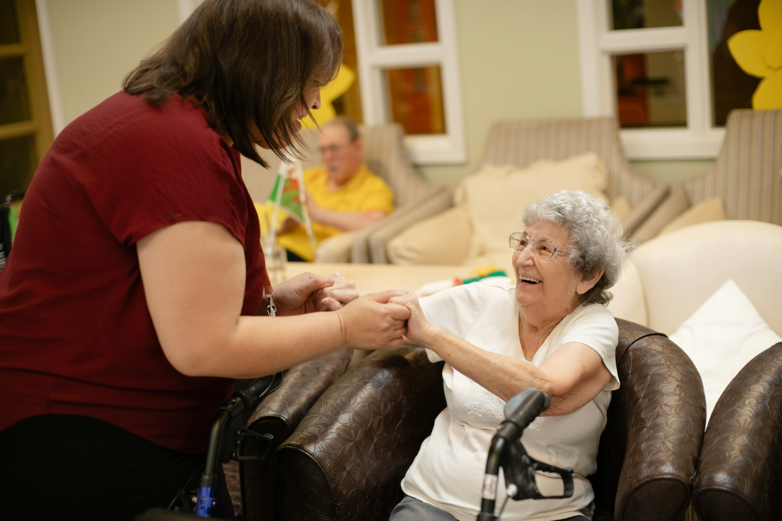 Carer holding hands with a smiling older woman seated in an armchair